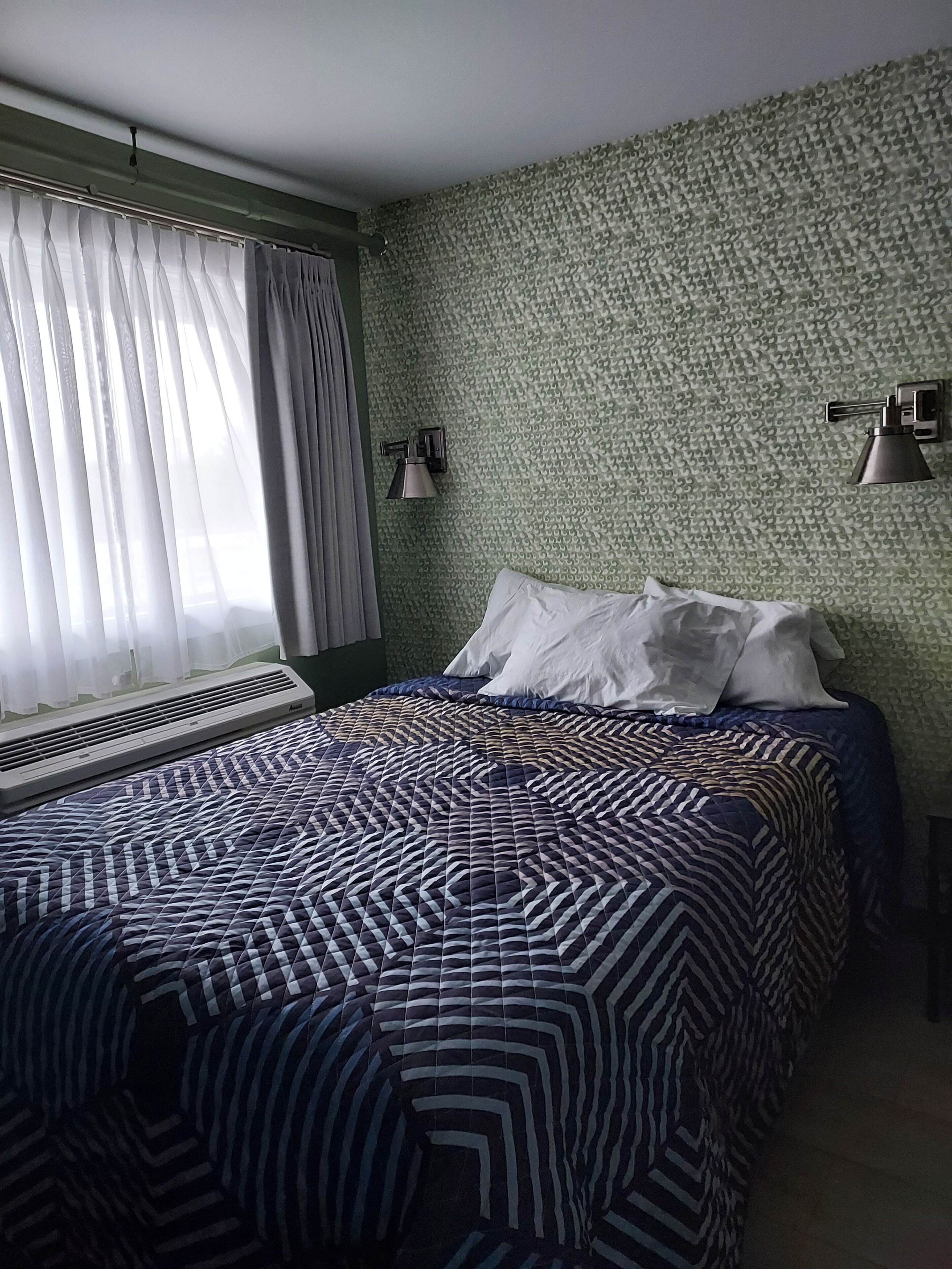 A bedroom with a patterned bedspread, a window with sheer curtains, and a wall-mounted lamp.