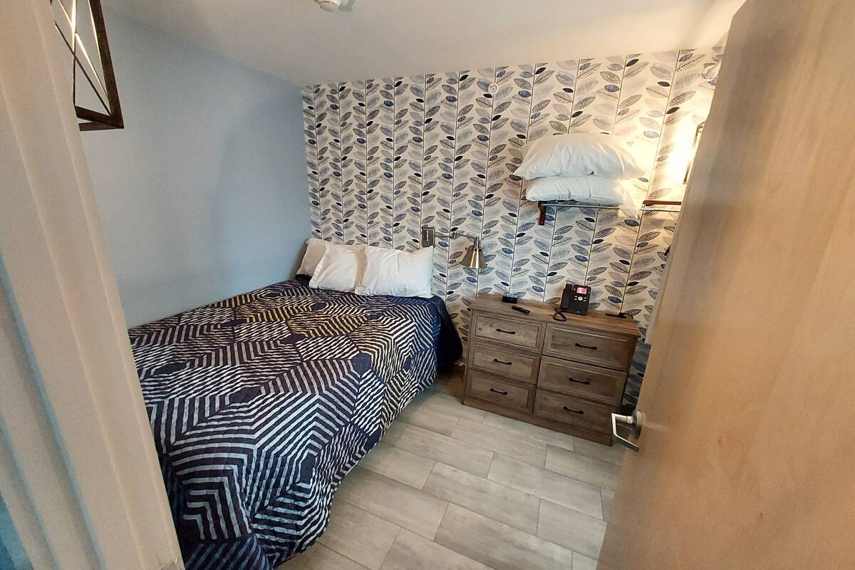 Un dormitorio con una colcha estampada, un aparador con teléfono y un papel tapiz decorado en la pared.