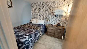 A bedroom with a patterned bedspread, a dresser with a telephone, and a patterned wallpaper on the wall.
