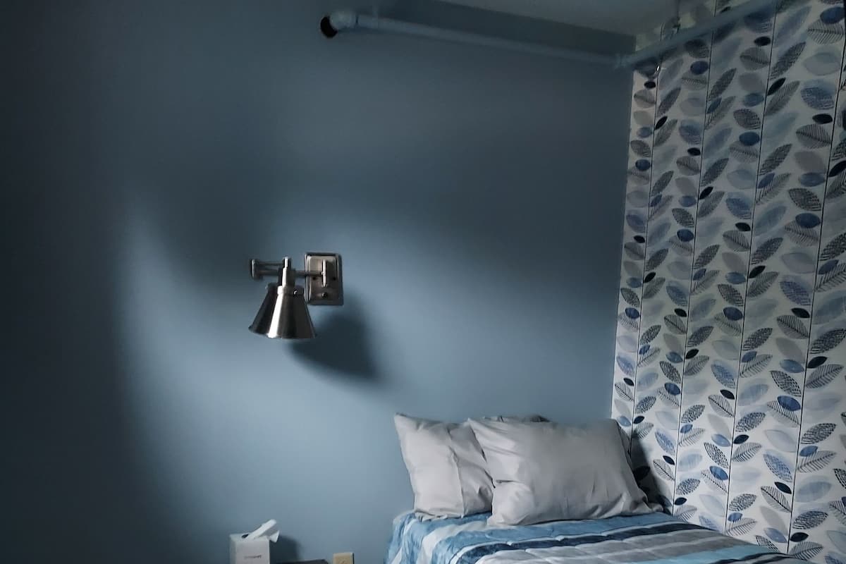 Un dormitorio con cama, mesita de noche, lámpara y cortina con estampado de hojas.