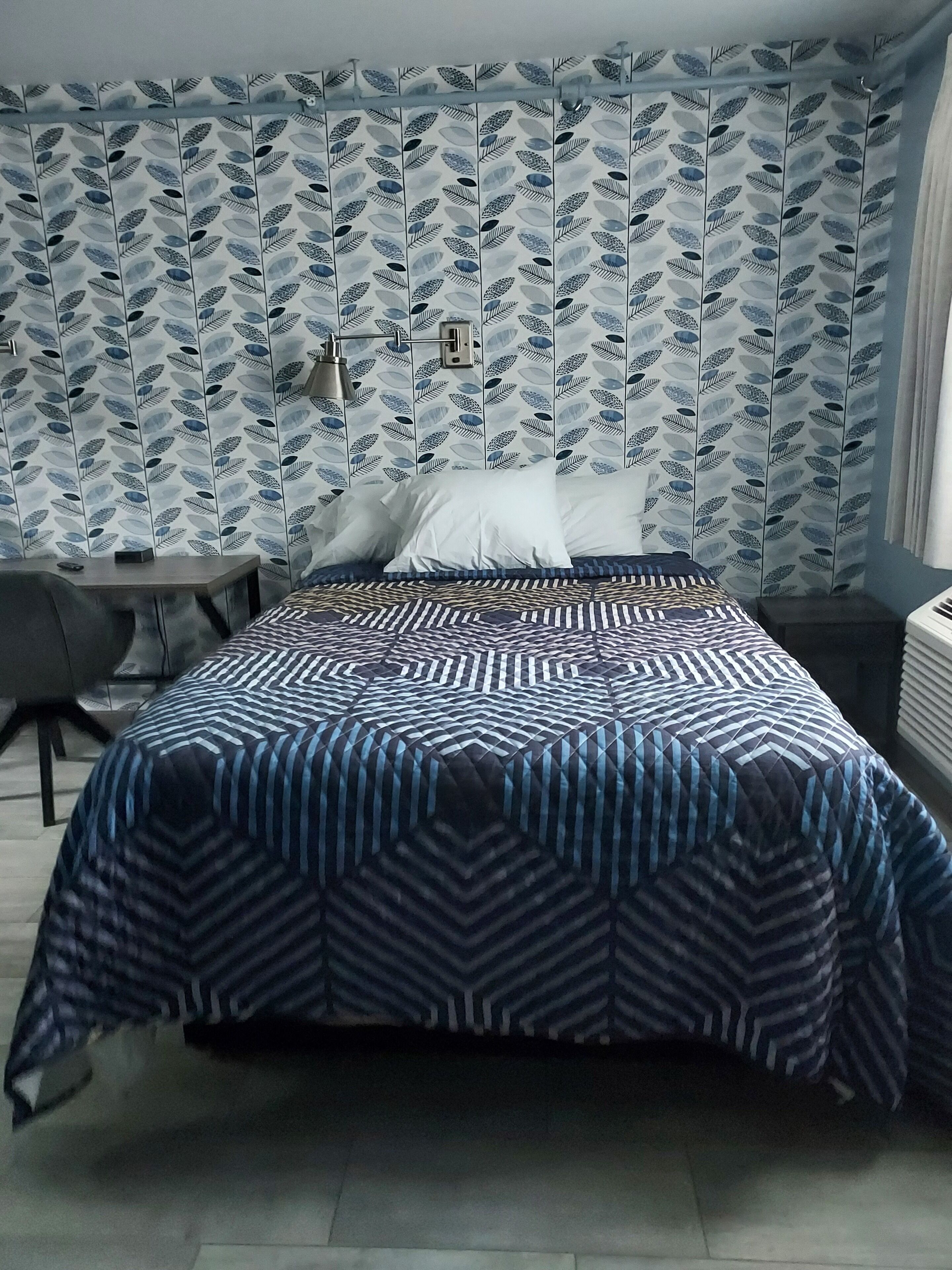 A bedroom with a patterned wallpaper, a bed with a geometric-patterned duvet, a desk, and a chair.