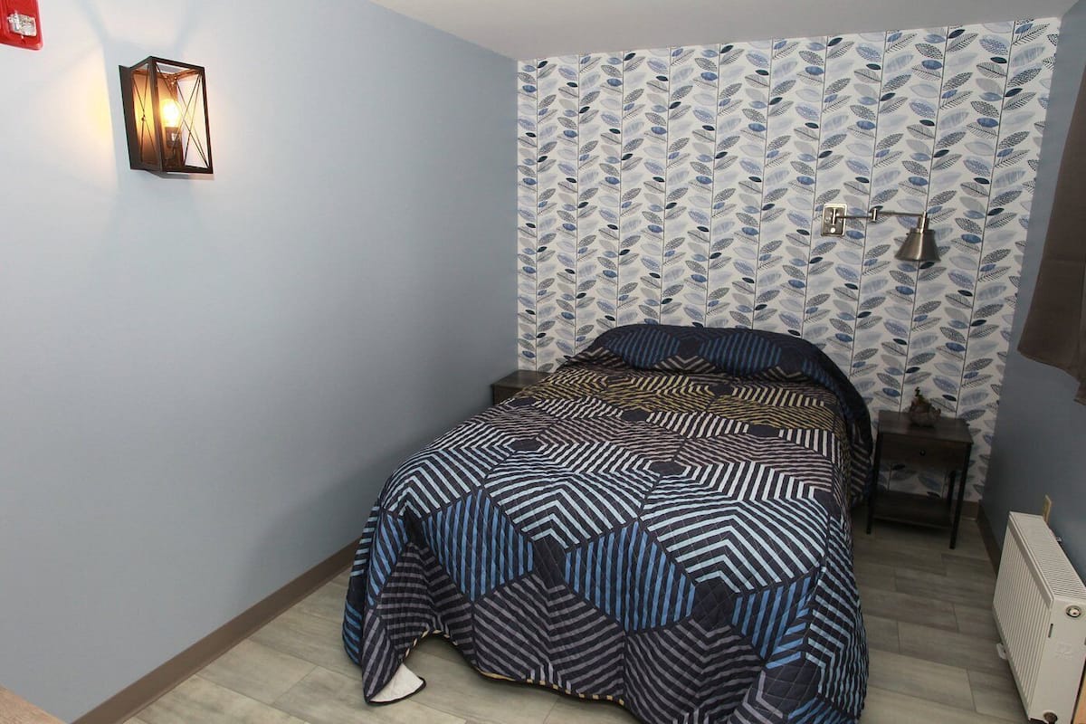 Un dormitorio con cama, luz empotrada en la pared, radiador y papel tapiz estampado.