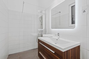 A modern bathroom with white tiles, a wooden vanity, a large mirror, and a shower. - Bright Apartment | Kitchen | Well Connected (Wien)