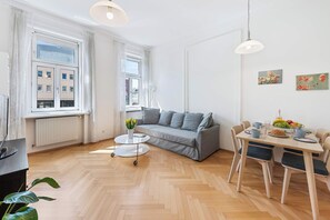A modern living room with a grey sofa, a dining table with chairs, a small round table with a vase of flowers, and large windows with curtains. - Bright Apartment | Kitchen | Well Connected (Wien)