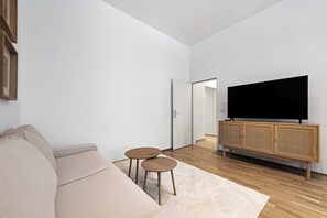 Ein modernes Wohnzimmer mit einer Couch, zwei kleinen runden Tischen, einem Fernseher auf einem Holzschrank und einer Tür zu einem anderen Raum.
