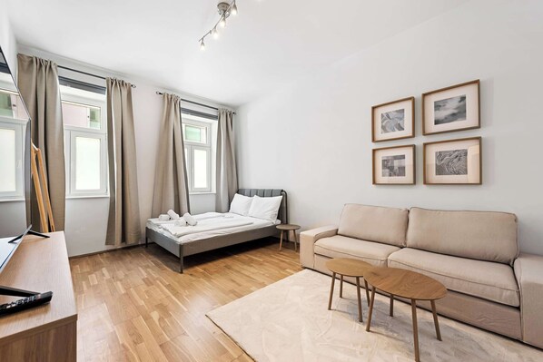 Ein modernes Schlafzimmer mit Bett, Sofa, Schreibtisch und einem kleinen Tisch. Vor den Fenstern hängen Vorhänge und an der Wand befindet sich ein Kunstwerk.