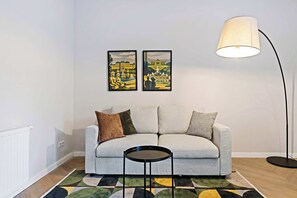 Un salon moderne avec un canapé gris, une petite table ronde, un lampadaire et deux tableaux encadrés représentant des paysages urbains accrochés au mur.