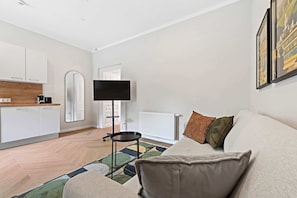 A modern living room with a sofa, a coffee table, a TV, and a kitchen area with cabinets and a radiator. - Kaiser Apartment | Comfort | Kitchen | Central (Wien)