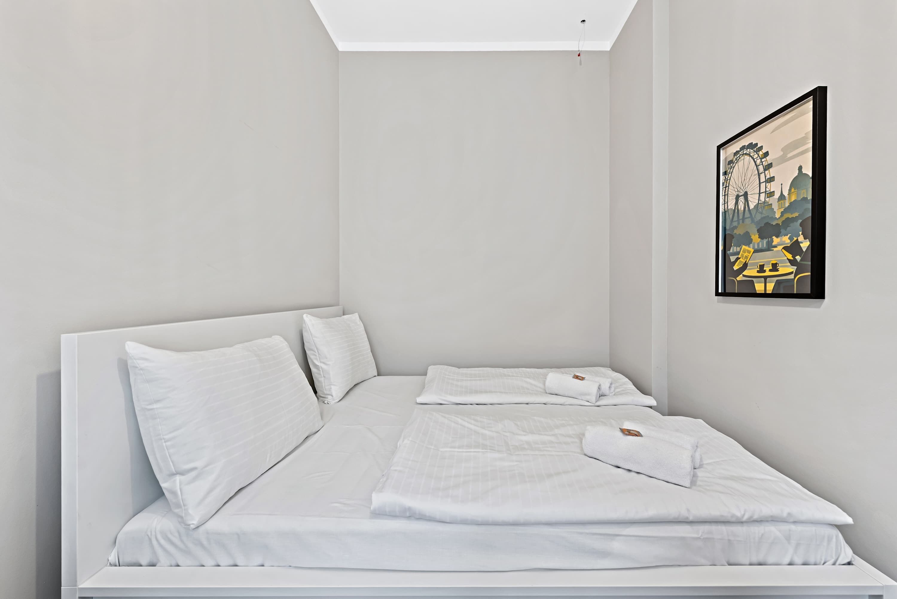 Un dormitorio minimalista con una cama individual, ropa de cama blanca y un cuadro con una vista urbana enmarcada en la pared.