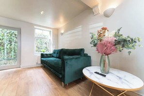 Ein Zimmer mit einem grünen Sofa, einem runden Tisch mit einer Vase und einem Fenster mit Blick auf Grünflächen.