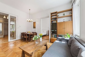 A living room with a wooden dining table, a sofa, a bookshelf, and a chandelier. - Stylish 3BR Apt. | Central | Kitchen | Balcony (Wien)