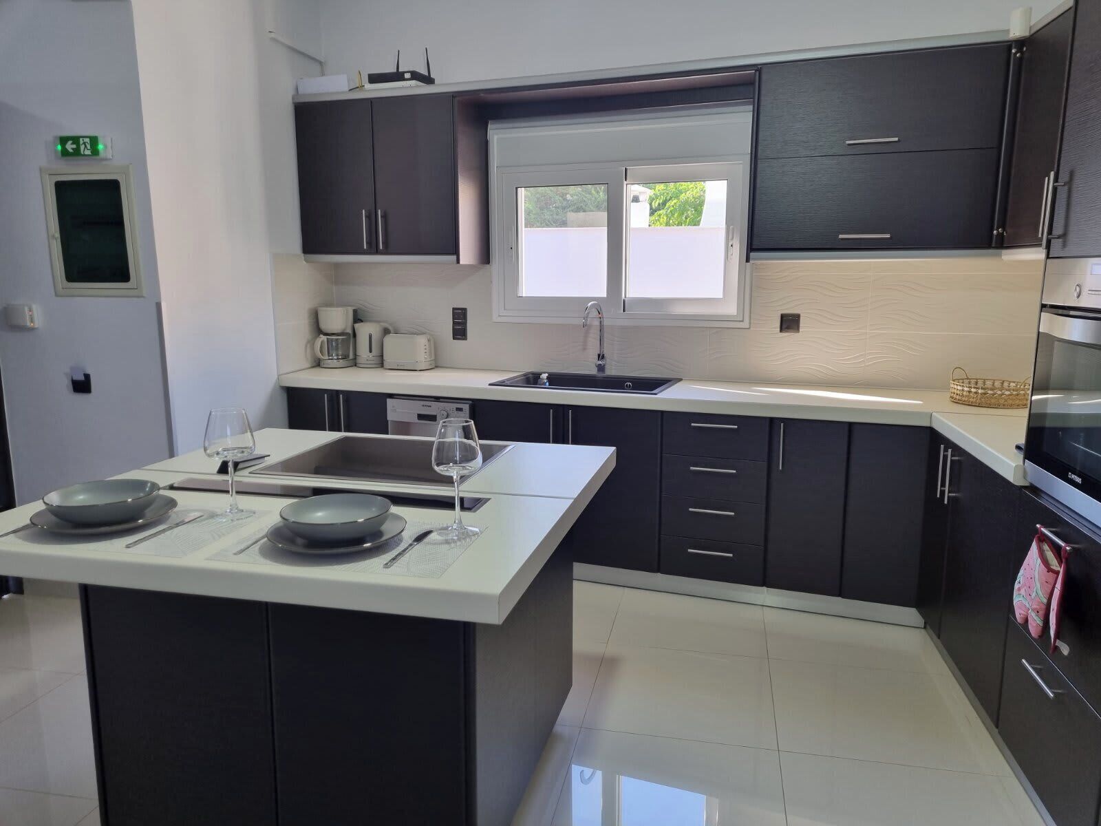 A modern kitchen with dark cabinets, a white countertop, a built-in oven, and a window providing natural light.