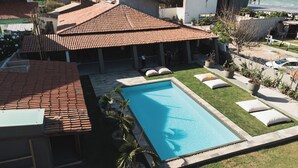 A swimming pool with a tiled roof, a grassy area with lounge chairs, and a view of neighboring buildings.