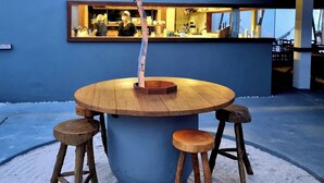 A round wooden table with stools, a tree, and a blue wall with a window.