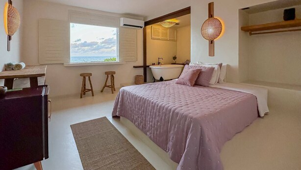 A bedroom with a bed, a desk, a window with a view, a bathtub, and a mirror.