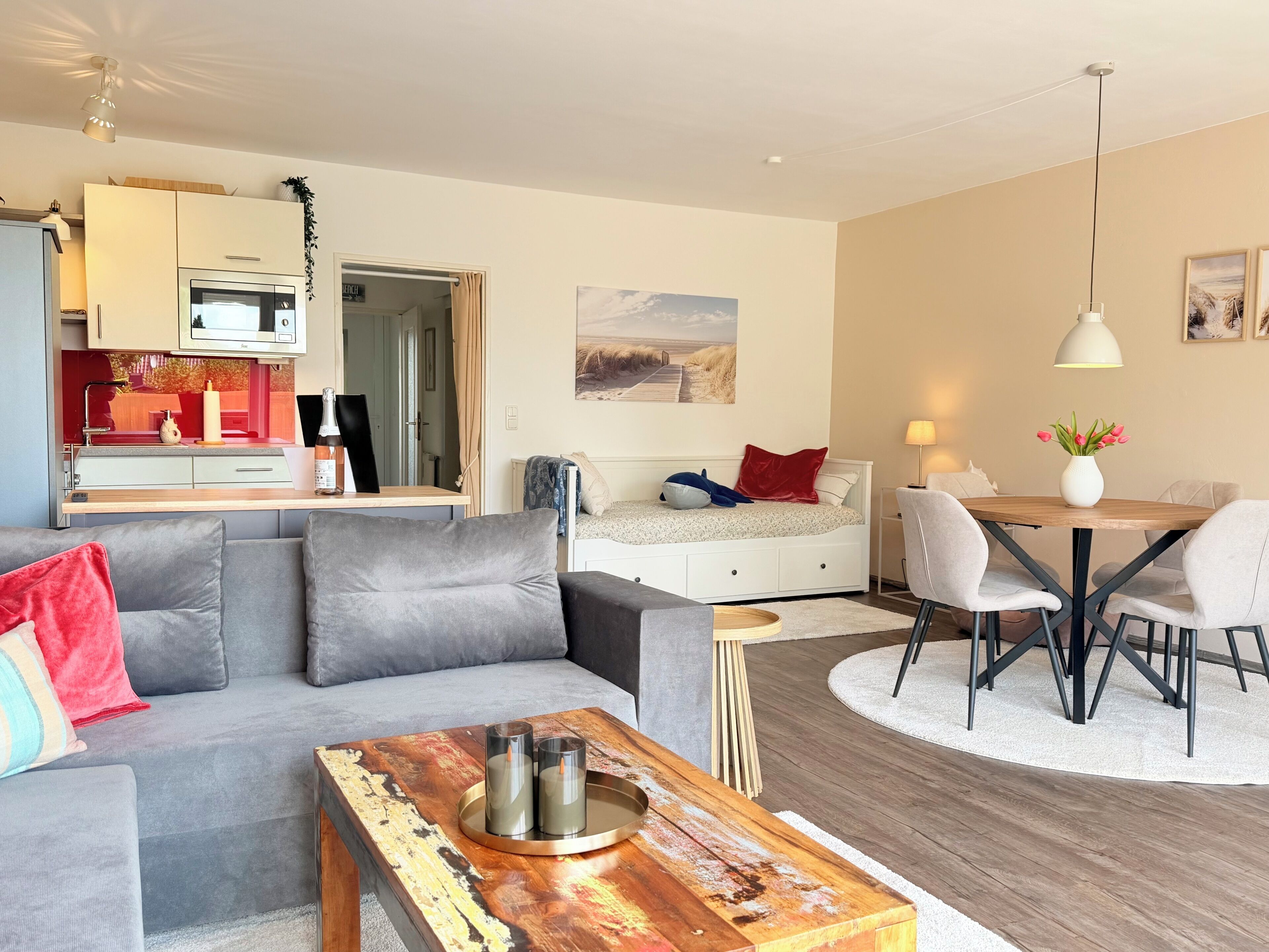 Una sala moderna con un sofá gris, una mesa de centro de madera con velas, una zona de comedor con una mesa y sillas blancas, y una cocina con microondas y armarios.