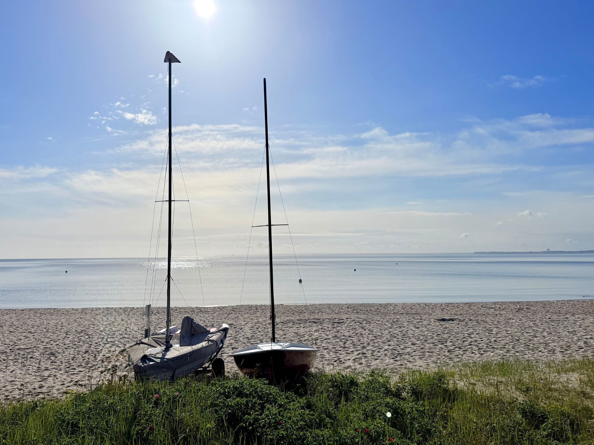 Ein Strand mit zwei Segelbooten, blauem Himmel und ruhigem Wasser.