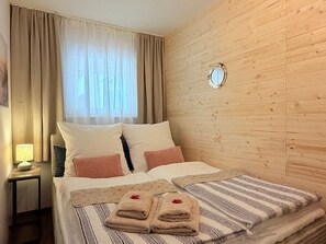 Una camera d'albergo con parete in legno, un letto con biancheria a righe e bianca, un comodino con una lampada e una finestra con tende trasparenti.