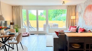 En moderne stue med spiseplads, en sofa, et sofabord, et fjernsyn og udsigt til en have gennem store skydedøre.