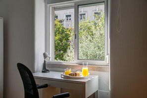 Ein kleiner Schreibtisch mit Stuhl, Lampe und Frühstücksbrett mit Croissants und Orangensaft neben einem Fenster.