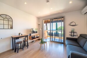 Ein modernes Wohnzimmer mit Sofa, Fernseher, Esstisch mit Hocker und Balkon mit Aussicht.