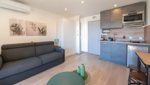 Ein modernes Wohnzimmer mit einer Couch, einem kleinen runden Tisch und einer Küchenzeile mit Schränken und Geräten.