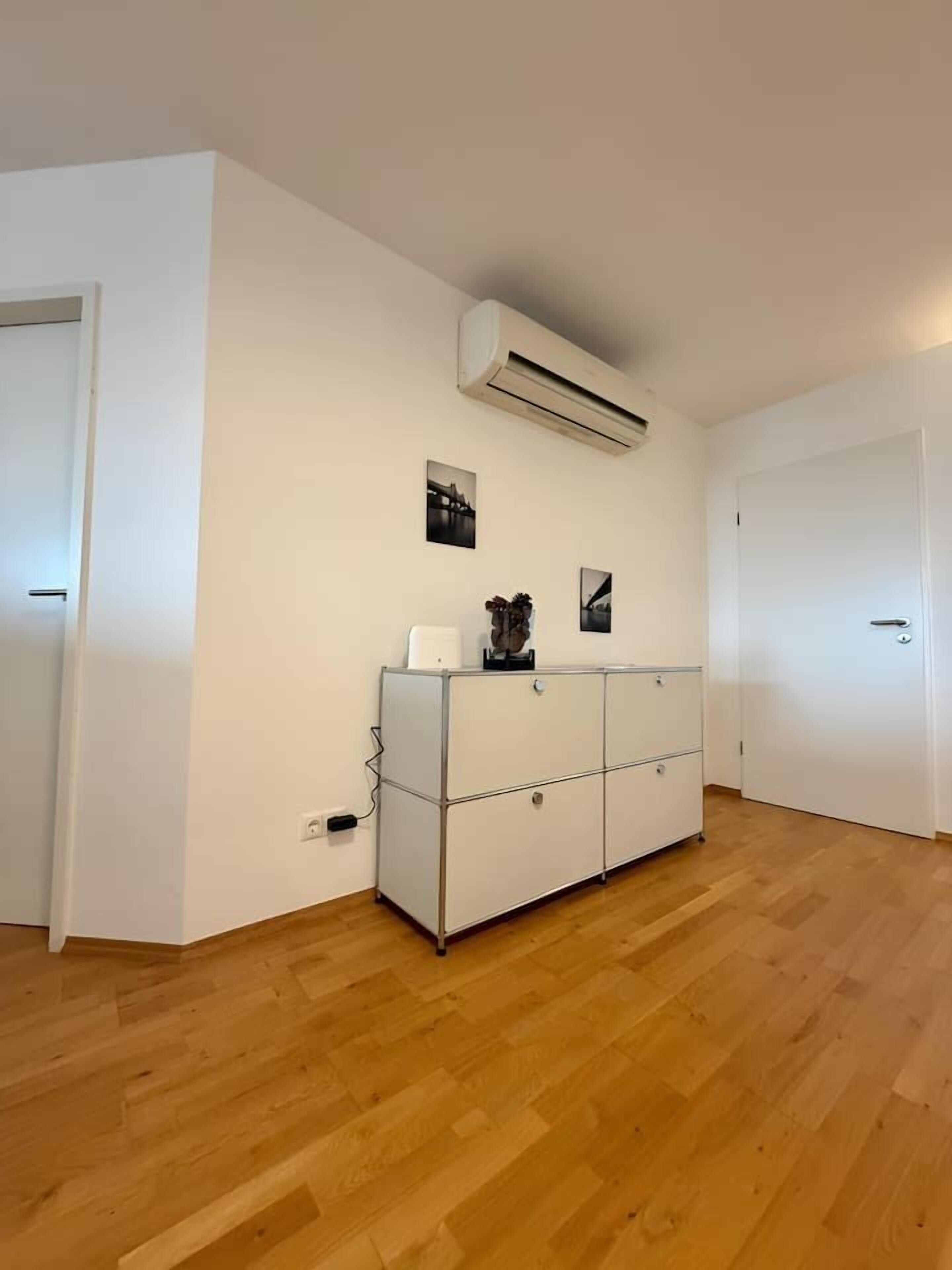 Een kamer met een houten vloer, witte wanden, een witte kledingkast en een airconditioning.