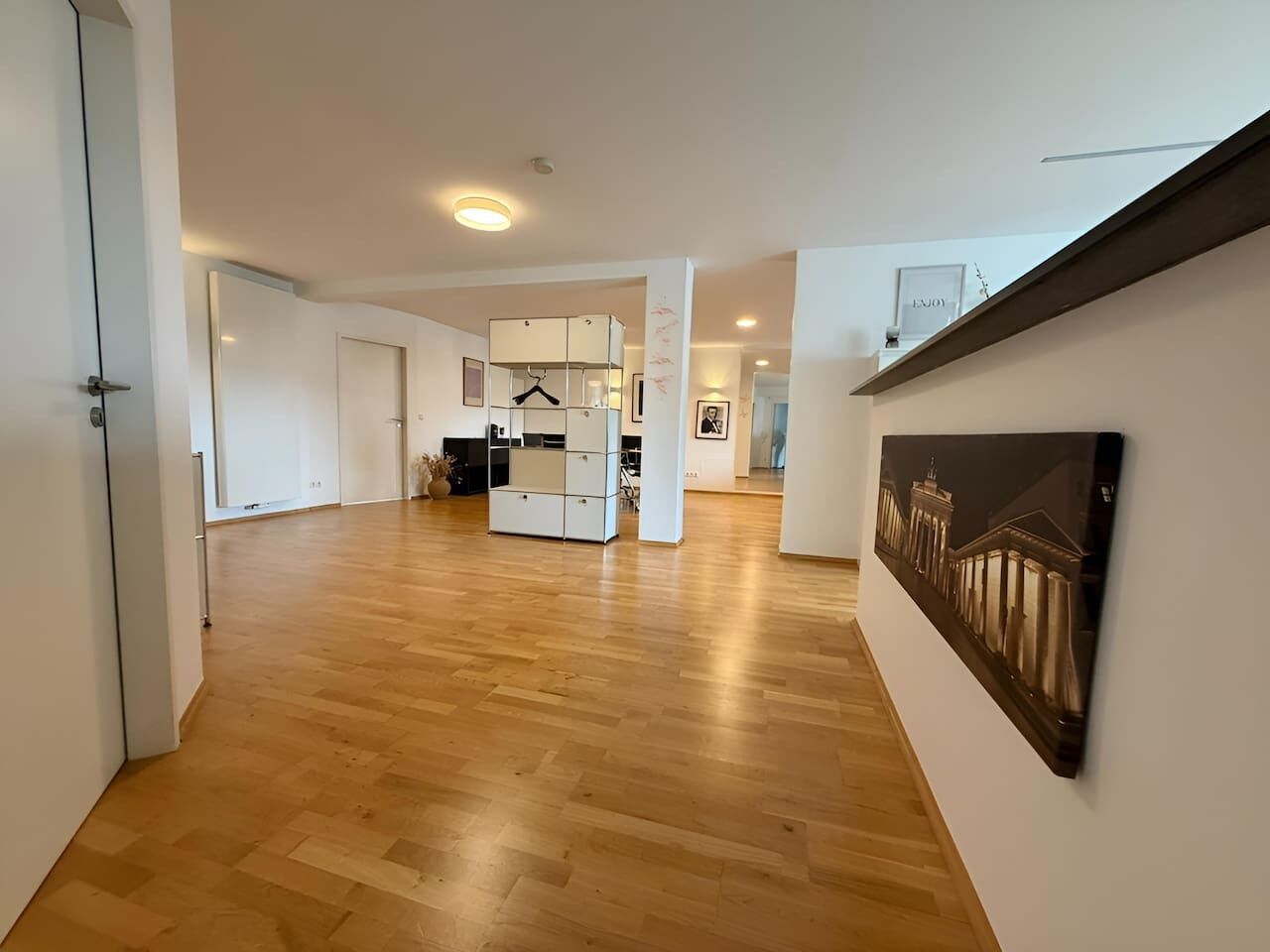A hallway with wooden flooring, a white wall with a framed picture, and a white cabinet with a hanging rack.
