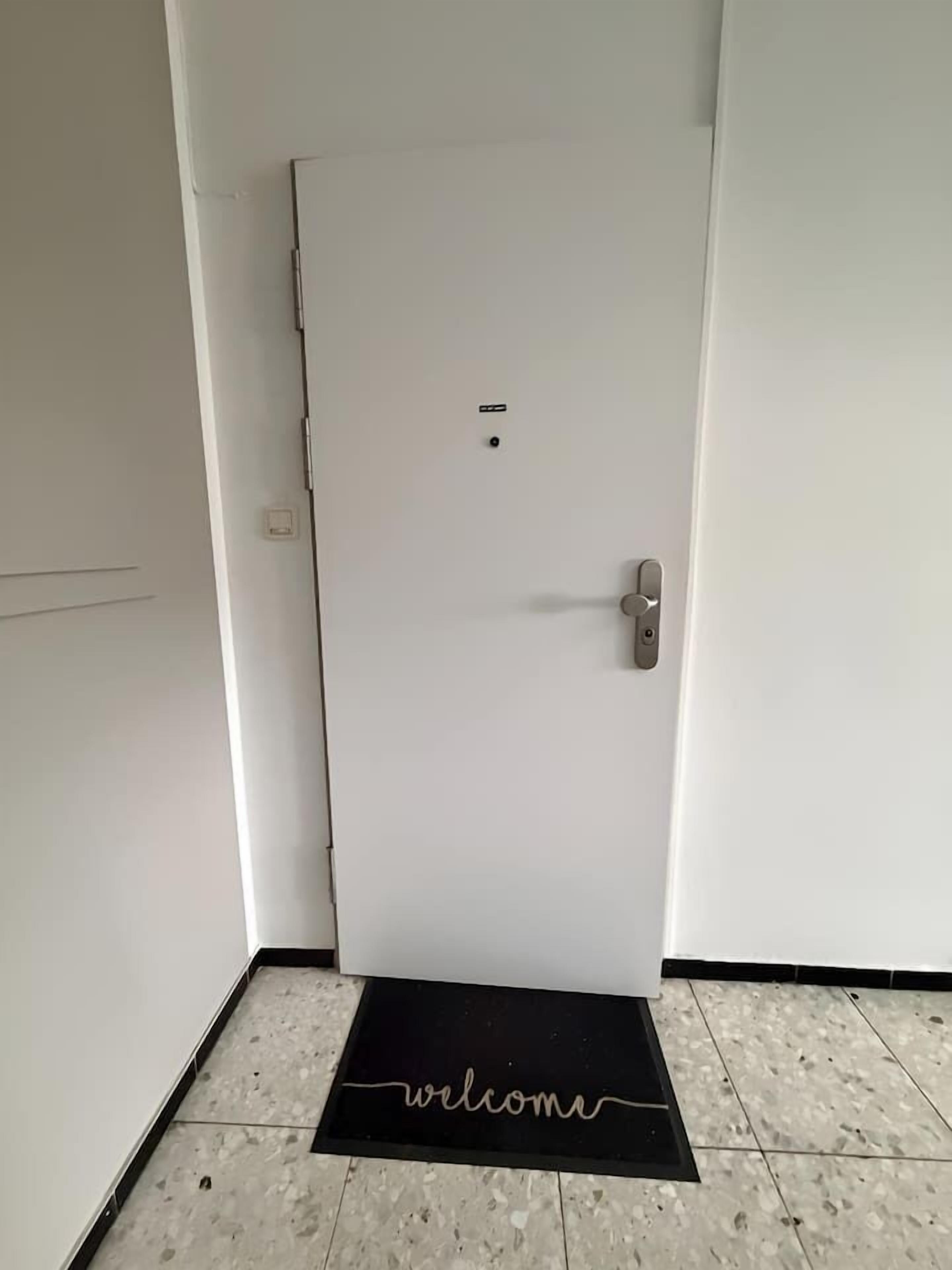 Une porte blanche fermée, dotée d’une poignée et d’une petite plaque de serrure, un paillasson noir sur lequel est inscrit le mot « bienvenue », et un sol carrelé.
