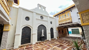 A courtyard with a white building featuring arched doorways and a statue, surrounded by stone walls and potted plants.