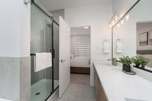 Une salle de bain moderne avec une douche à parois de verre, un meuble-lavabo blanc avec un miroir, et un lit en arrière-plan.