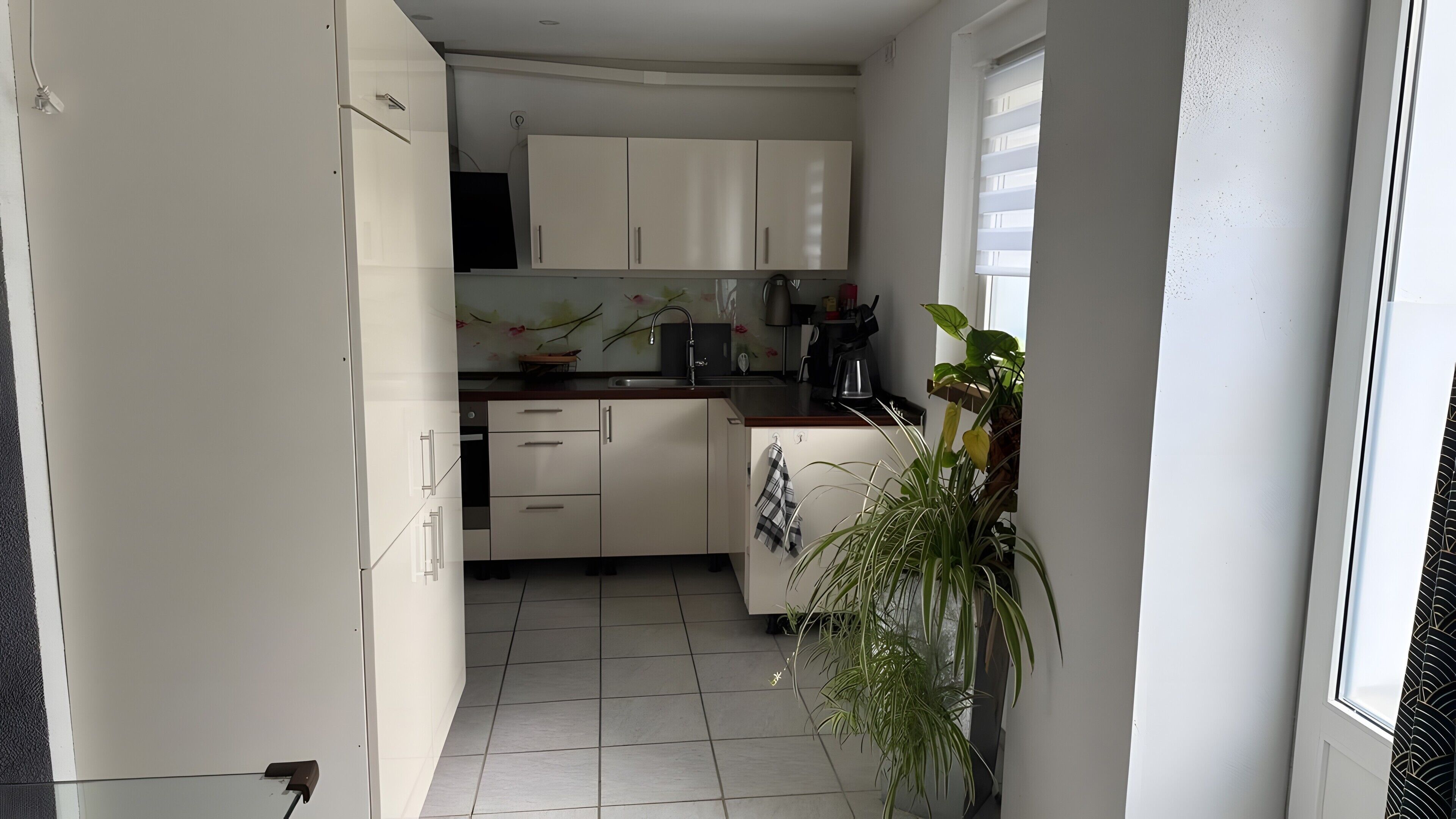 Una cocina estrecha con armarios blancos, piso de baldosas y una ventana con persianas.