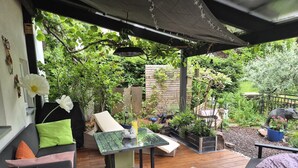 Un pati amb una taula de vidre, un sofà i una cadira. Hi ha un jardí amb plantes diverses i una estàtua.
