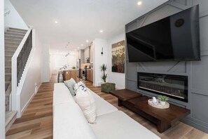 A modern living room with a large flat-screen TV, a fireplace, a sofa, and a staircase. - 3BRHome Near Rogers Place & NAIT w/ Garage Parking (Edmonton)