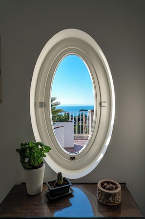 Ein ovales Fenster mit Meerblick, ein Blumentopf und ein kleiner Kaktus auf einem Tisch.