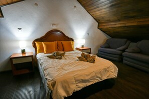Quarto com teto de madeira, cama com edredom branco, duas luminárias de cabeceira e um sofá cinzento.
