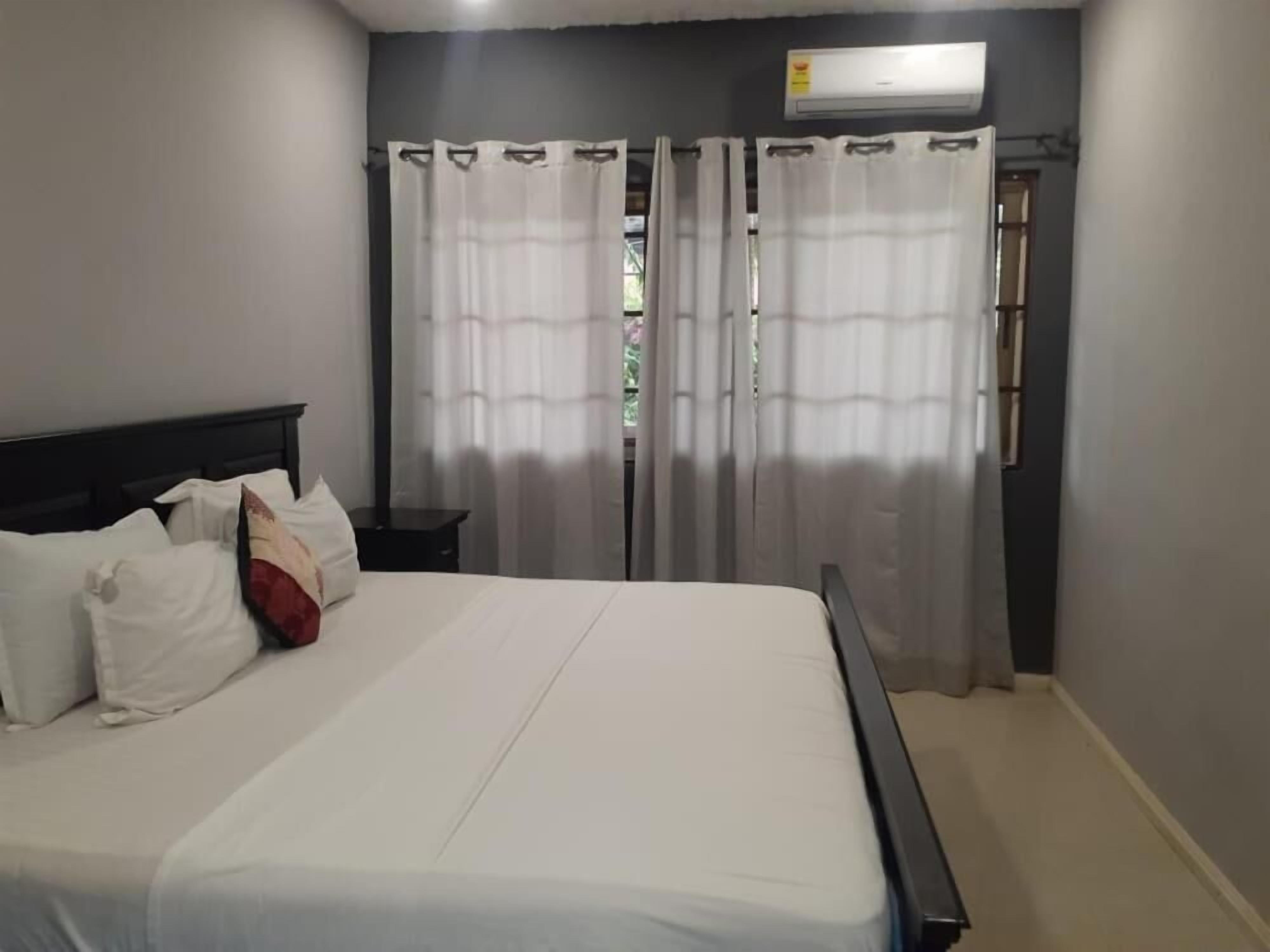 Un dormitorio con cama, cortinas blancas, una mesita de noche y un aire acondicionado instalado en la pared.