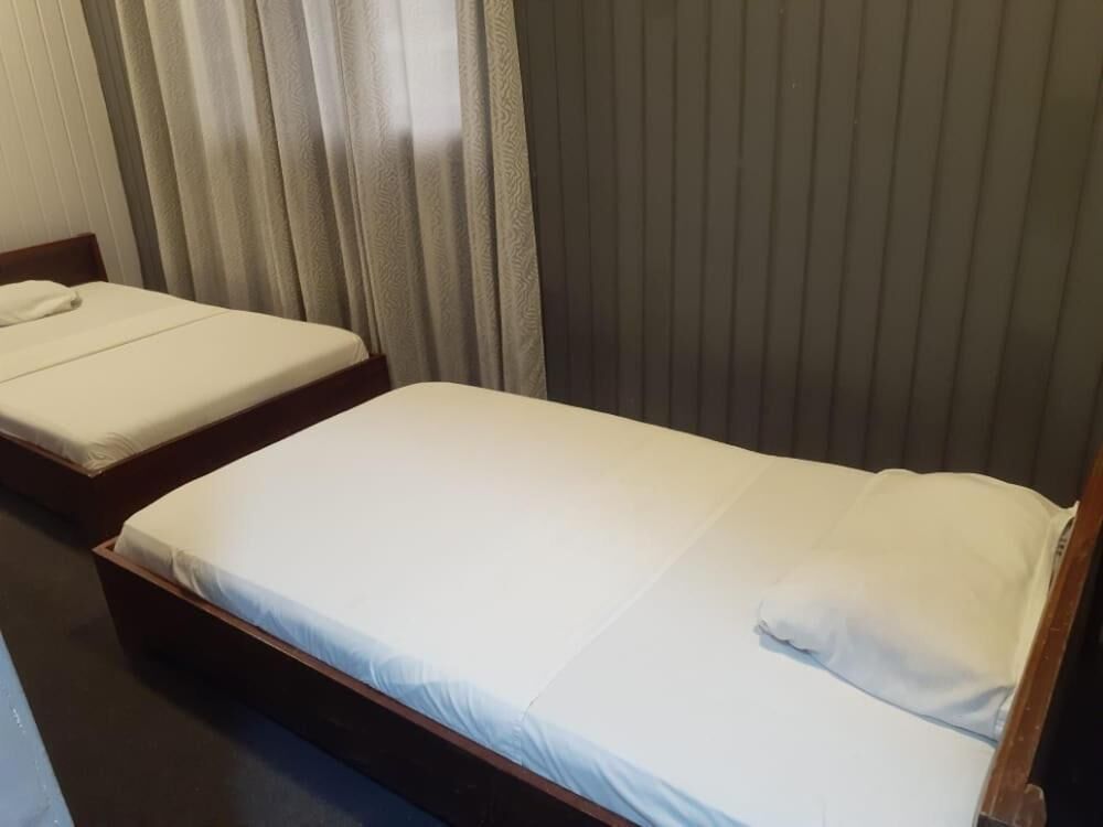 A single bed with a wooden frame and white bedding in a room with a dark-colored wall and a window with vertical blinds.