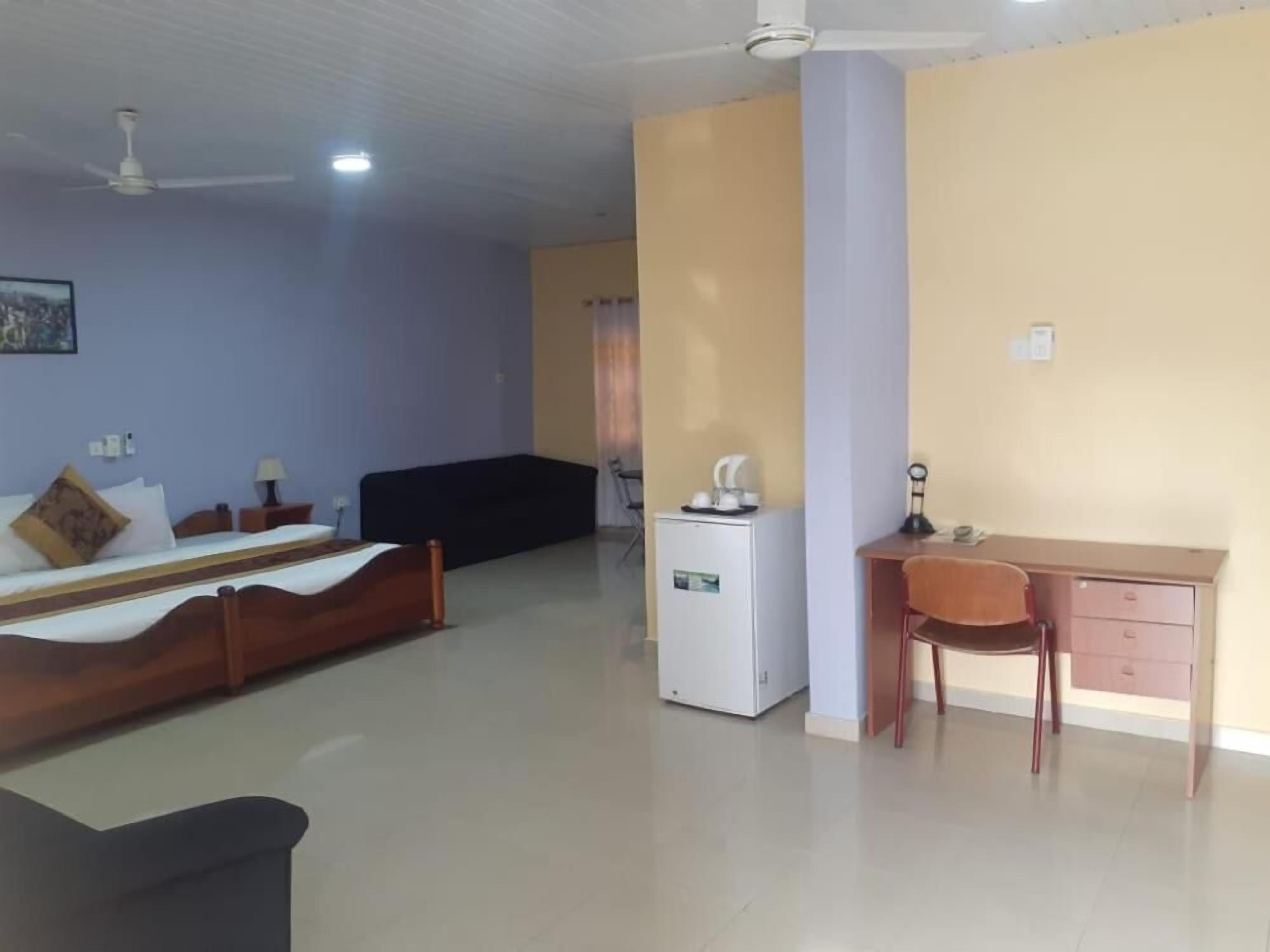 A room with a bed, a desk, a chair, a refrigerator, and a ceiling fan.