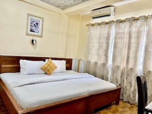 A neatly made bed with white bedding, a wooden headboard, a wall-mounted air conditioner, and a patterned curtain. - Hans Cottage Botel (Cape Coast)