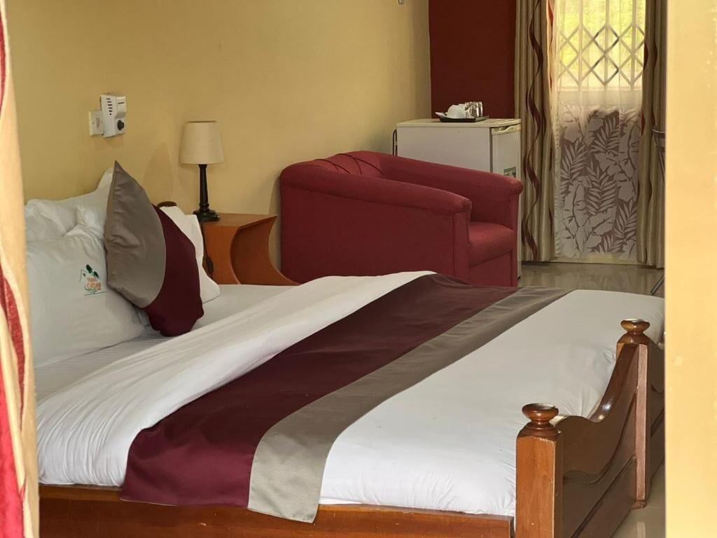 A hotel room with a bed, a red armchair, a small table with a lamp, a white cabinet, and a window with curtains.