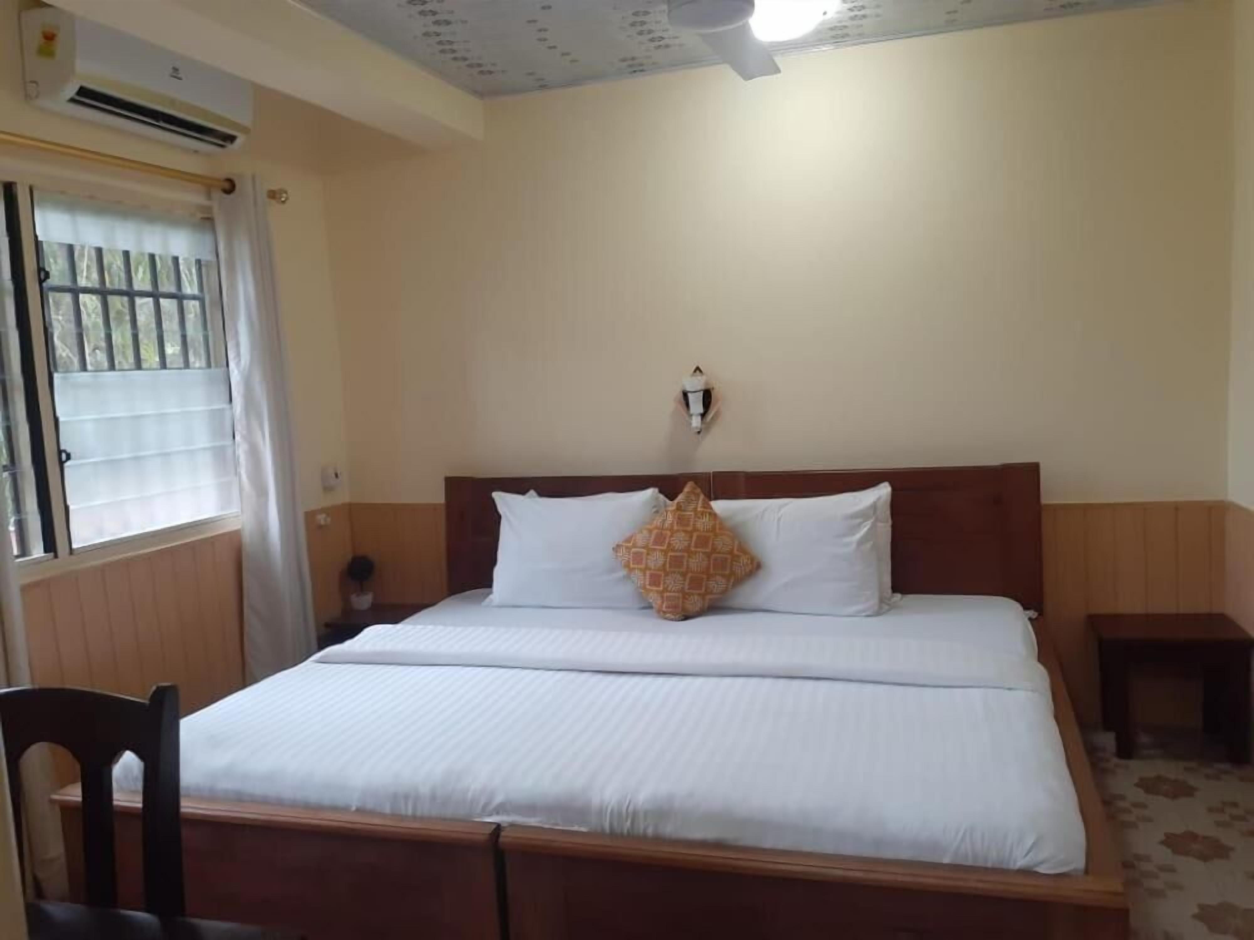 Una habitación de hotel con cama, mesita de noche, silla, ventana con cortinas y aire acondicionado.