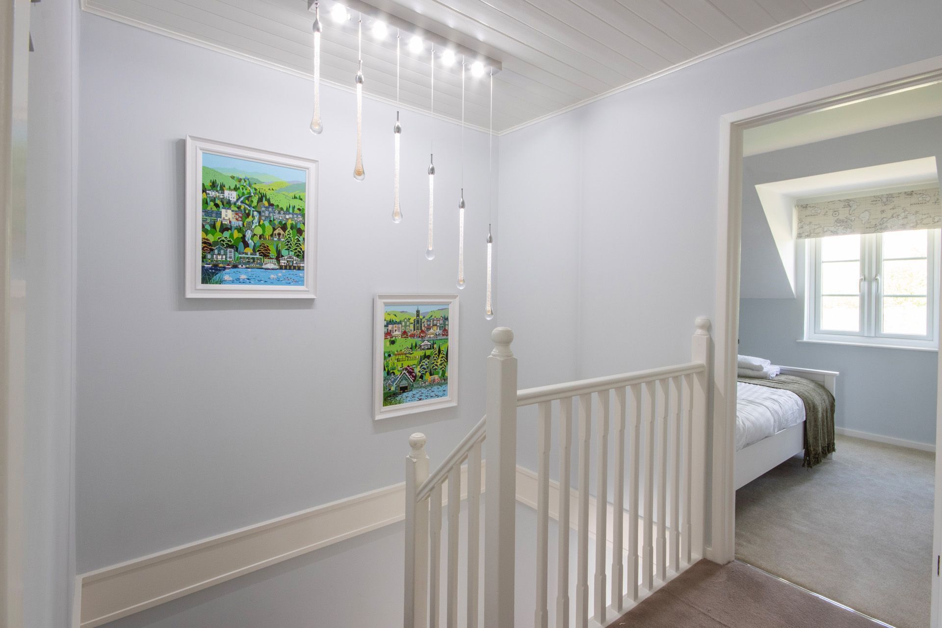 A bedroom with a bed, a window with curtains, a staircase with a railing, and two framed pictures on the wall.