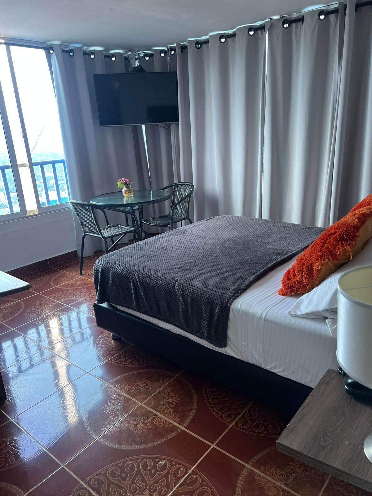 Una habitación de hotel con cama, una mesa con sillas, televisor y balcón con cortinas.