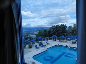 Zona de piscina con sombrillas azules y sillones de descanso, vista a un paisaje urbano y una piscina con diseño azul.