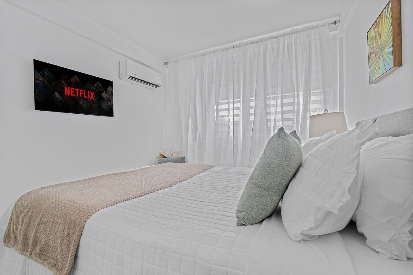 A bedroom with a bed, a blanket, pillows, a wall-mounted TV, and a window with curtains.
