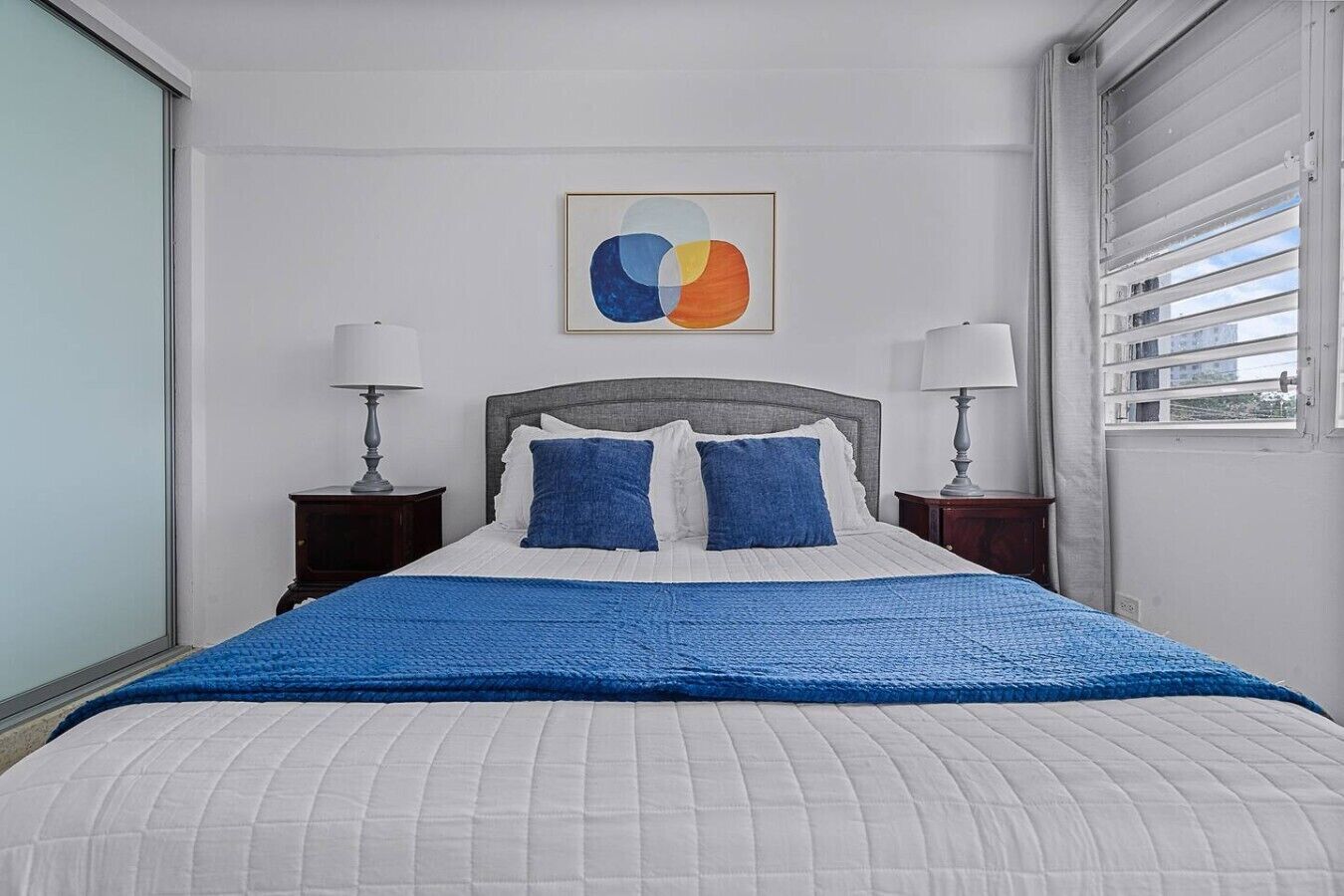 A bed with a blue blanket, two pillows, a headboard, two bedside tables with lamps, and a wall art piece.