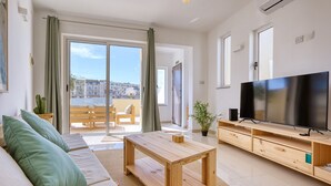 A modern living room with a sofa, a wooden coffee table, a TV, and a balcony with a view of buildings. - Charming bright penthouse with 3 Terraces JCUT2-1 (Swieqi)