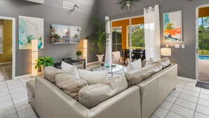 Amplia sala de estar con un sofá seccional beige grande, una mesa de centro y un comedor con mesa y sillas. Hay dos ventanas grandes que dejan entrar luz natural y ofrecen vistas al exterior.