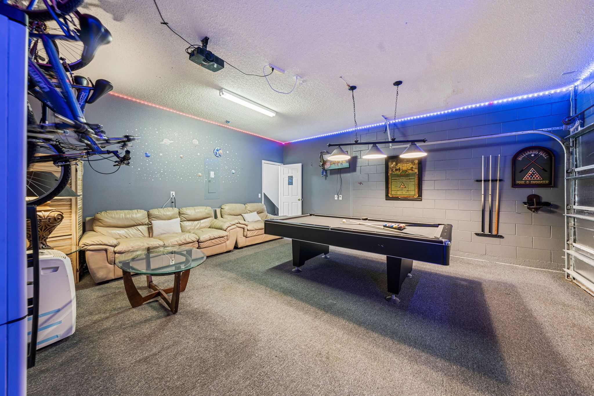 Una habitación con mesa de billar, sofá, proyector instalado en el techo y cartel en la pared.